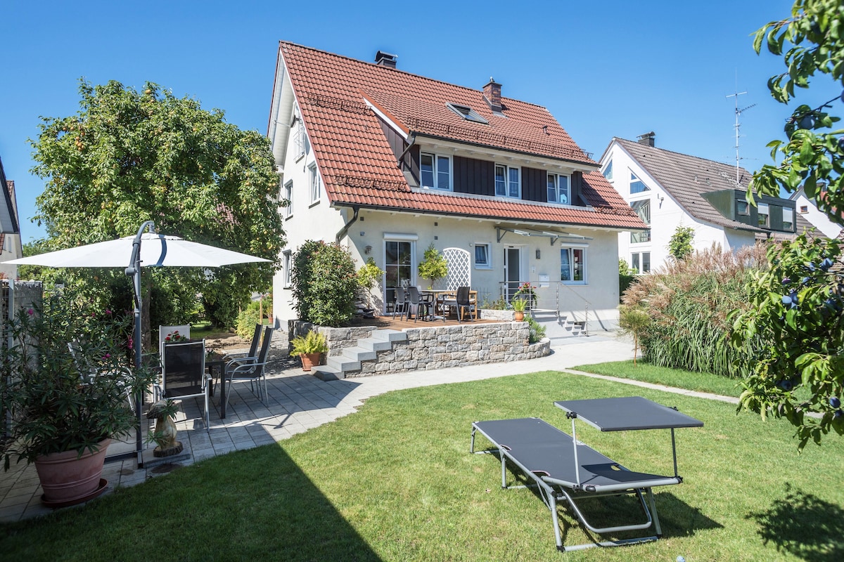 The exterior of the vacation apartment shows a multi-level house with a red-tiled roof. A well-kept garden surrounds the property, featuring a sunbathing area with loungers and a shaded seating arrangement. Greenery enhances the inviting outdoor space, providing a relaxing environment.