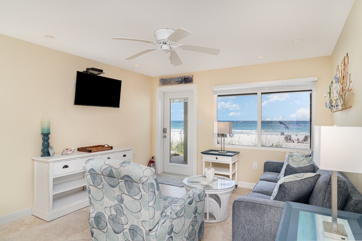 The living area is furnished with comfortable seating arrangements, including a patterned armchair and a grey sofa. A round glass table sits in the center, while a flat-screen television is mounted on the wall. Natural light floods the space from the sliding glass door, which leads to the beach.