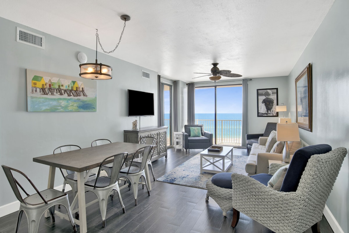A spacious living area features large glass doors opening to a private balcony with ocean views. A dining table with metal chairs is positioned beside a decorative console. Comfortable seating arrangements include various chairs and a sofa, with artwork adorning the walls.