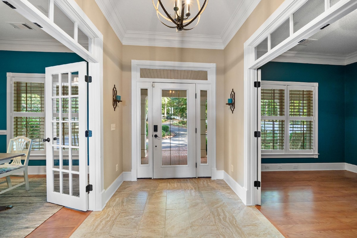 A welcoming entrance is shown, featuring double glass-paneled doors framed by stylish wall décor. Natural light fills the space, highlighting the warm wood flooring and decorative lighting fixture overhead. Light teal walls complement the inviting atmosphere.