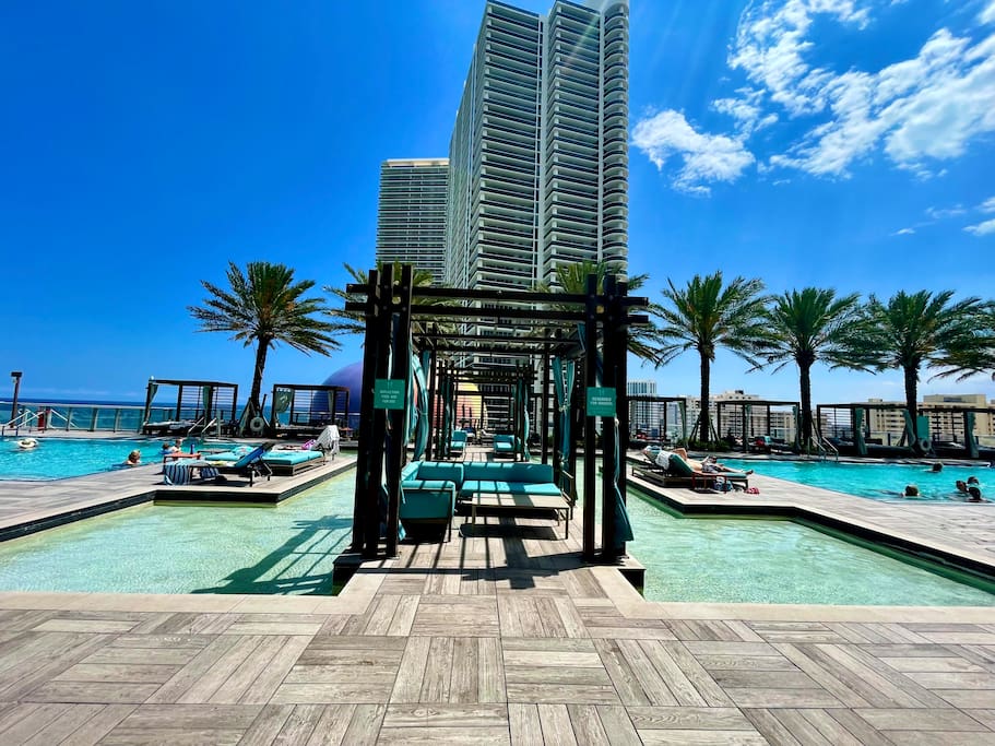 Two infinity pools are visible, surrounded by wooden decks and palm trees. Comfortable lounge chairs are arranged along the pool area. The sky is clear with a few clouds, and the tall resort building is seen in the background, providing a vibrant atmosphere for relaxation.