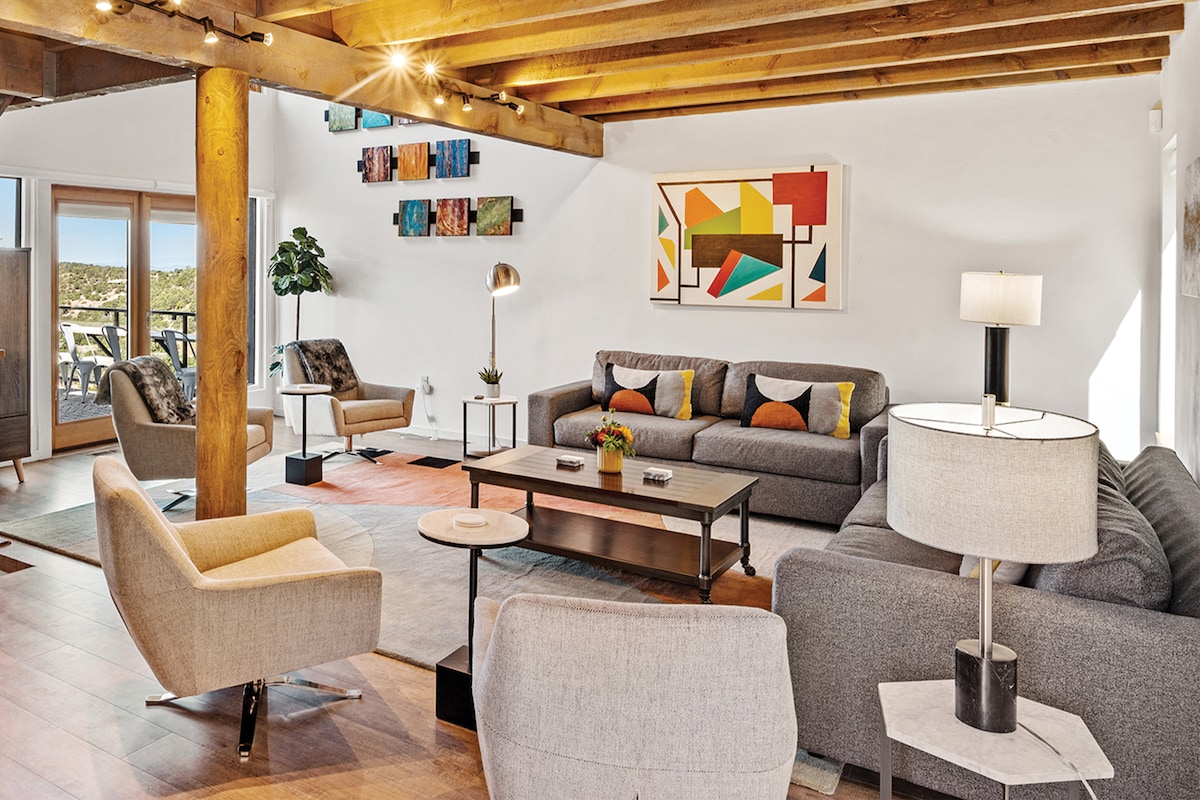 A spacious living area is designed with modern furnishings, featuring multiple seating options including gray sofas and stylish armchairs. Large windows provide ample natural light and views of the outdoors. Colorful artwork and decorative accents add a vibrant touch to the room.