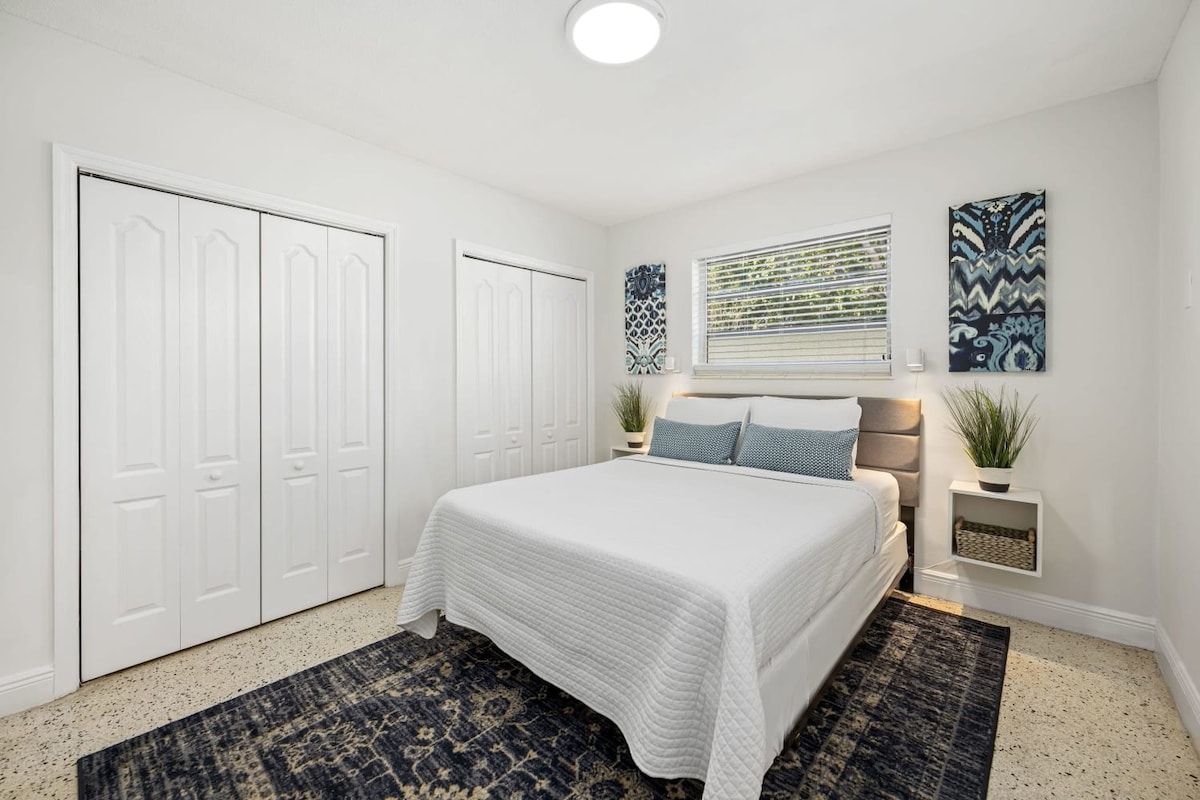 A bright, minimalist bedroom features a spacious bed with light bedding and two accent pillows. Decorative wall art complements the neutral walls, while potted plants add a touch of greenery. A large window allows natural light to fill the room, enhancing the open feel.