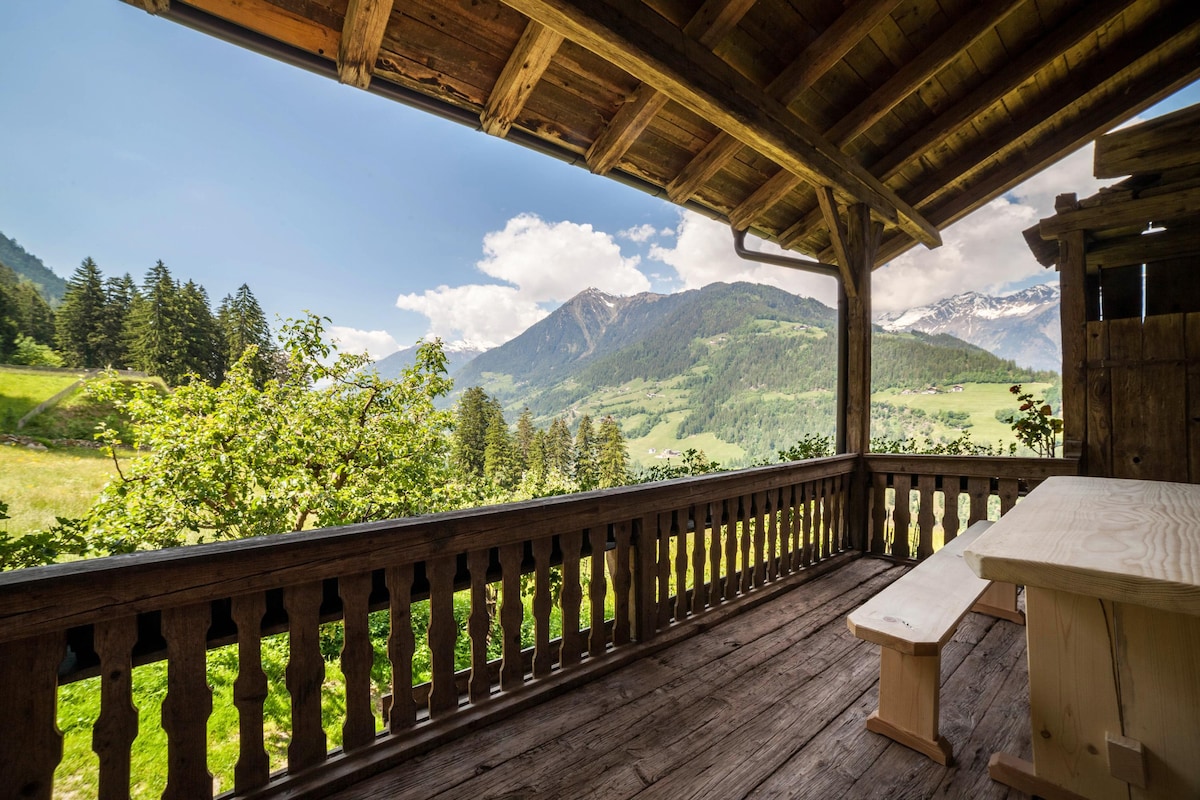 A covered terrace features wooden seating and a table, offering panoramic views of the surrounding mountains and valleys. The rustic wooden railing complements the natural scenery, which includes lush greenery and distant peaks, enhancing the outdoor experience.
