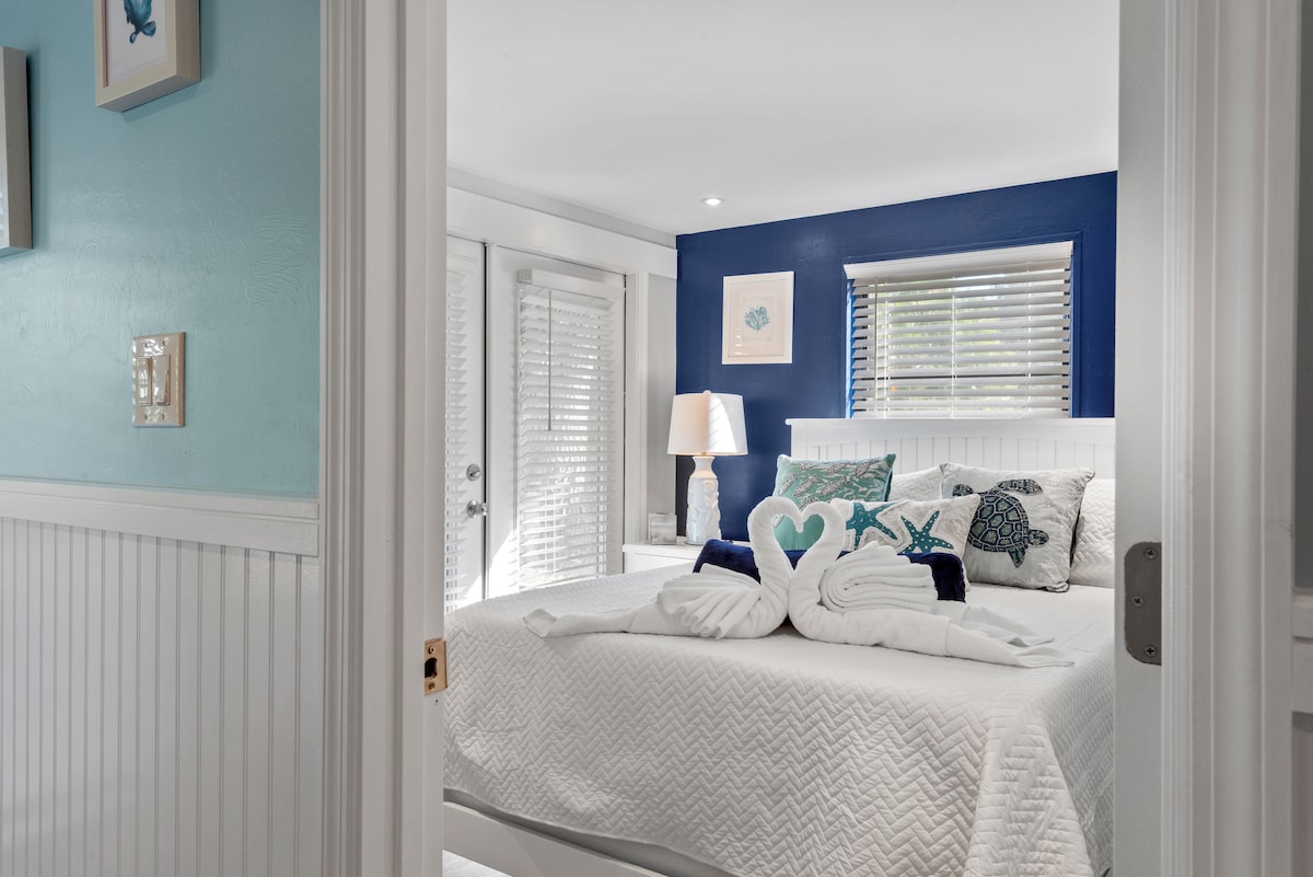 A serene bedroom is featured, showcasing a spacious bed adorned with light-colored linens and decorative pillows in coastal hues. A lamp on a nightstand casts gentle light, with sliding glass doors allowing natural light. The walls are painted in soothing shades of blue and white.