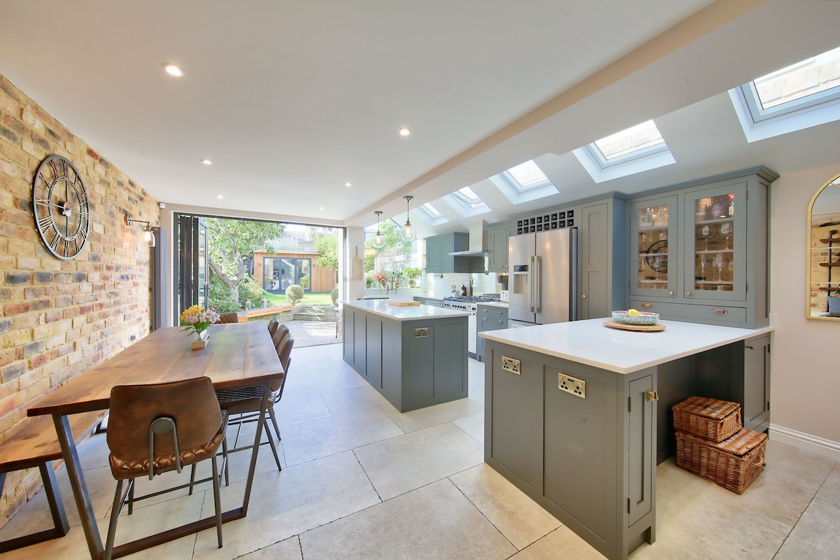 The kitchen area showcases a spacious layout featuring a central island, large dining table, and modern appliances. Natural light floods the room through skylights and glass doors, which lead to the garden. A brick wall adds texture to the contemporary design.