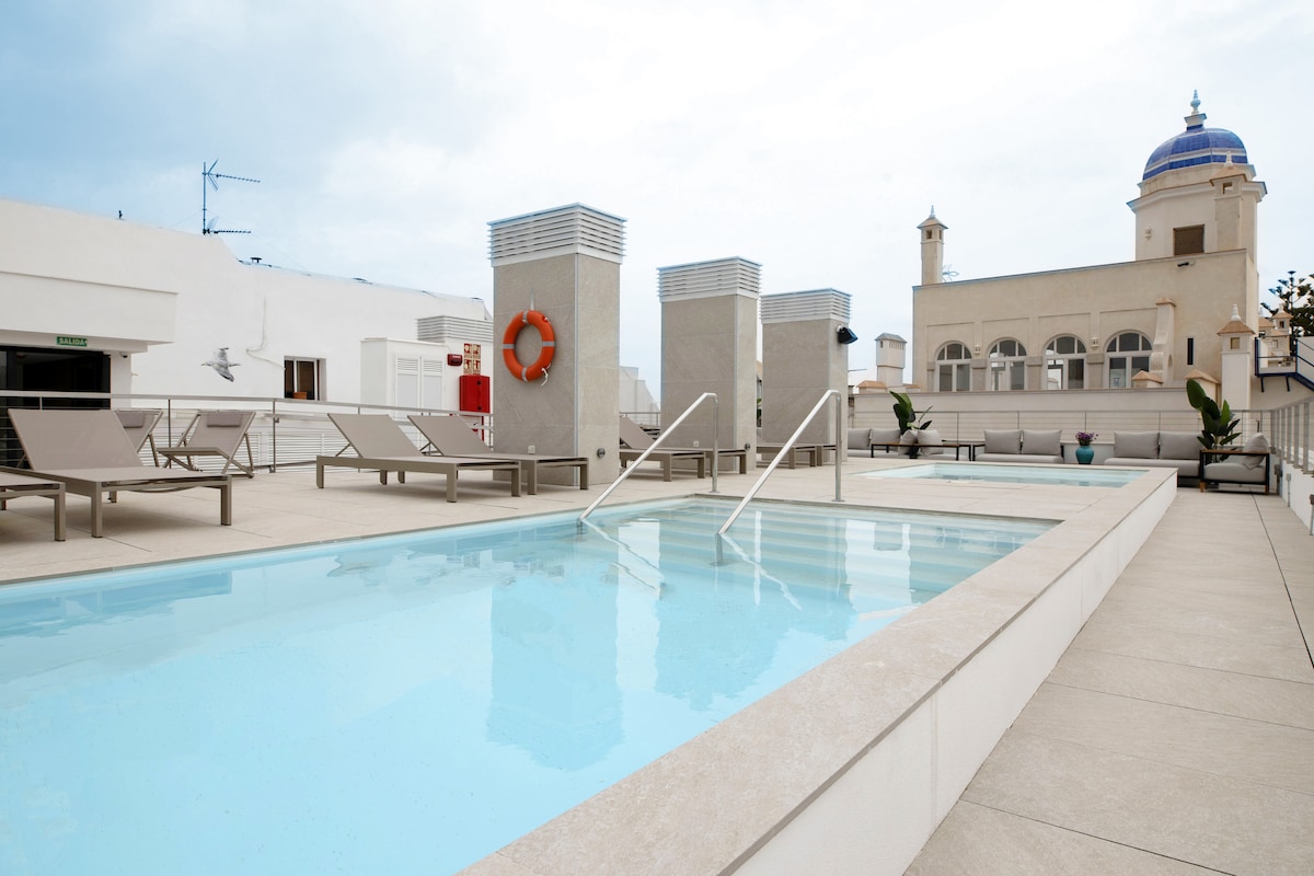 A rooftop terrace features a clear swimming pool with gentle steps leading in. Sun loungers are positioned around the pool area, complemented by safety elements such as a life preserver mounted on the wall. The skyline includes a notable building with a dome in the background.