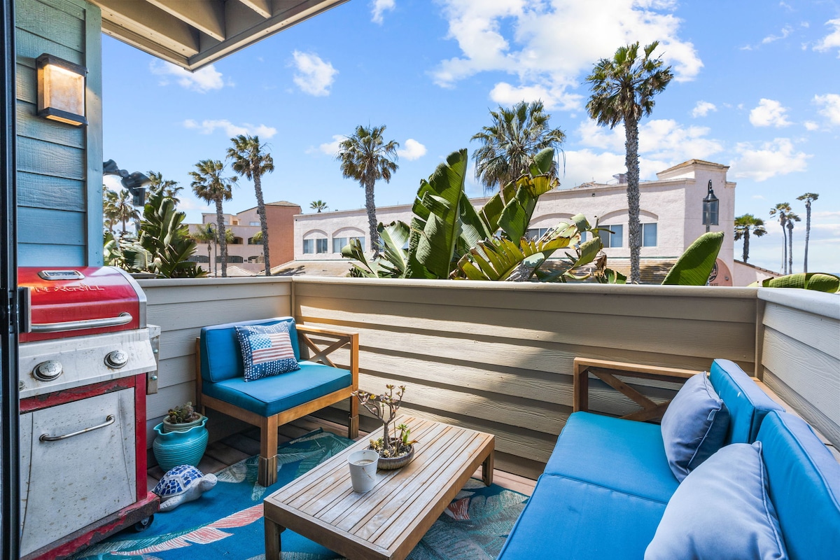 A cozy outdoor seating area offers blue cushions on wooden furniture, positioned on a balcony surrounded by palm trees and lush greenery. A grill is nearby, with a small table and potted plant complementing the space. Bright clouds and a hint of blue sky create a light atmosphere.
