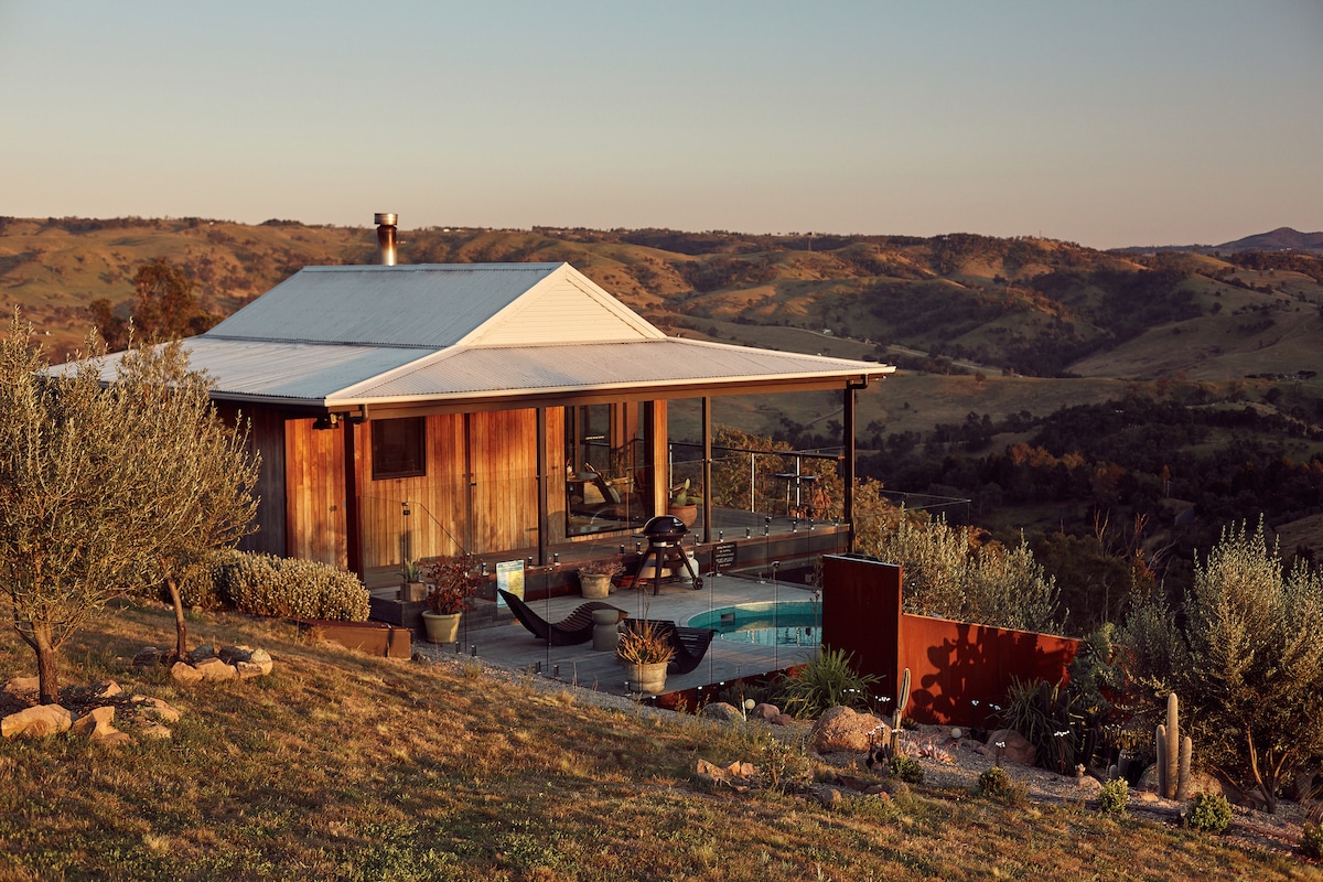 A rustic cabin set against rolling hills during golden hour. The structure features a large covered terrace, surrounded by lush greenery and potted plants. A plunge pool is visible beside a cargo net hammock, inviting relaxation amidst the expansive landscape.