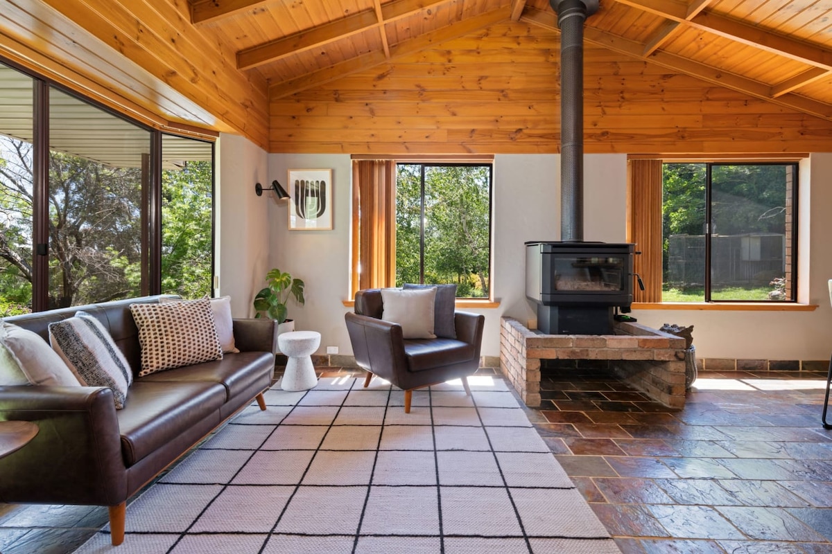 A spacious living area features a cozy wood-burning stove, complemented by wooden ceiling beams. Comfortable seating arrangements include a dark sofa and armchairs, positioned around a neutral area rug. Large windows allow natural light to illuminate the room and provide views of the surrounding greenery.