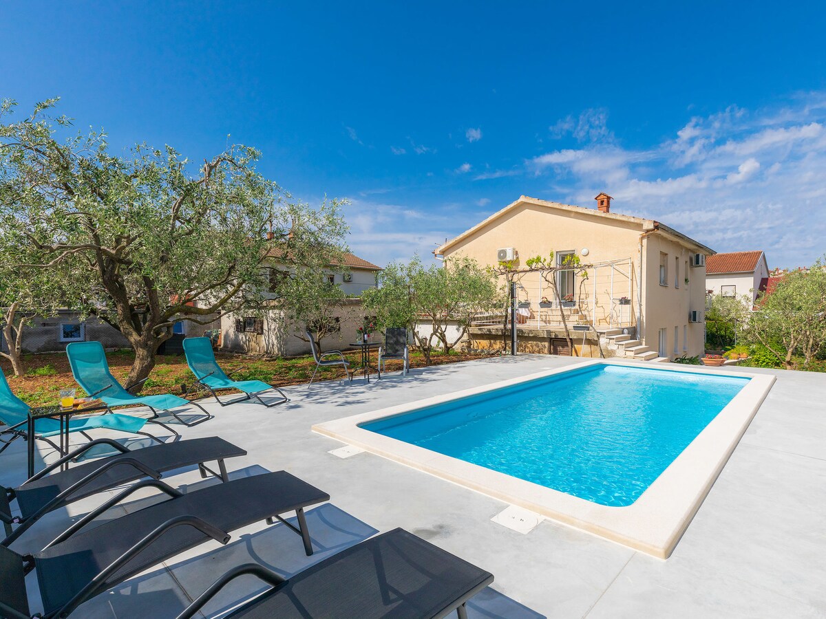 A swimming pool is situated within a spacious outdoor area, surrounded by olive trees. Sun loungers are set along the poolside. A light-colored two-family house is visible in the background, under a clear blue sky with scattered clouds.