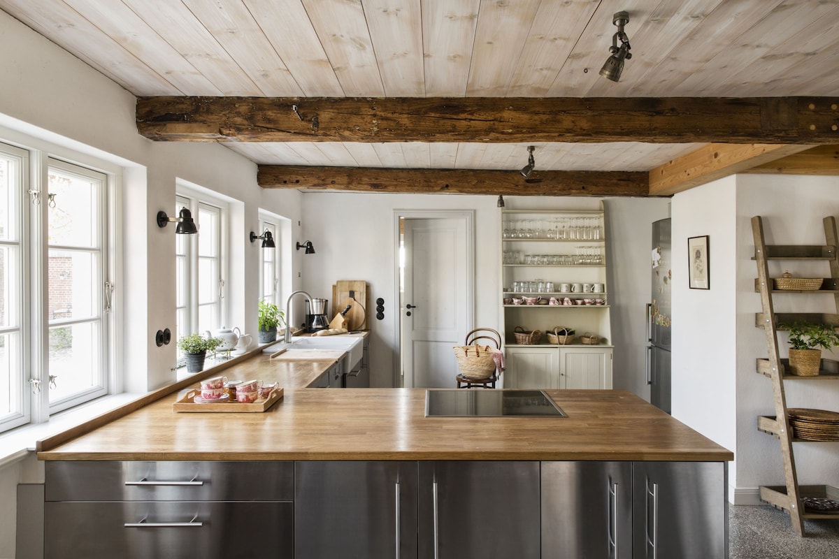 A spacious kitchen features a central wooden island with stainless steel cabinetry. Large windows allow natural light, illuminating the room's rustic wooden beams and modern appliances. Shelves with glassware and plants add charm, while a door leads to a connected space.