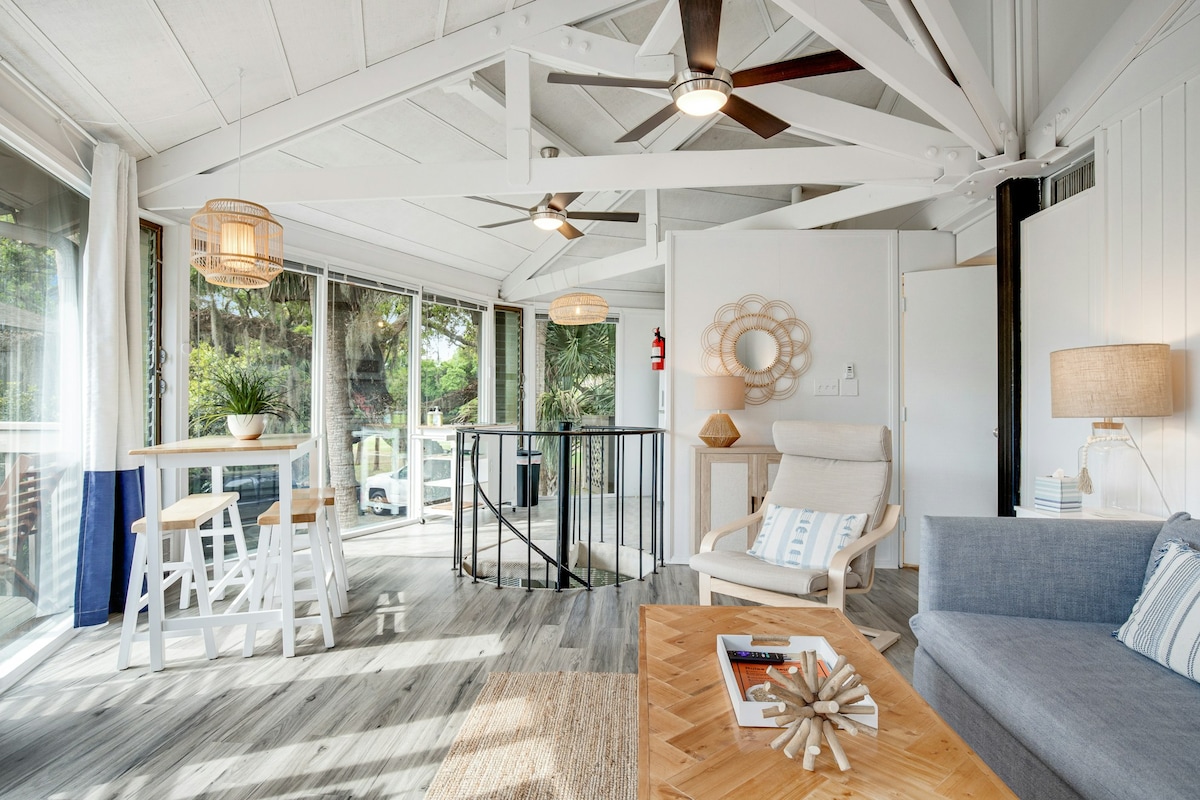 The airy living room features wood beam ceilings and large windows that allow natural light to fill the space. Comfortable furnishings include a gray sofa and a light-colored armchair, complemented by a round decorative mirror and a coffee table with a natural wood finish.