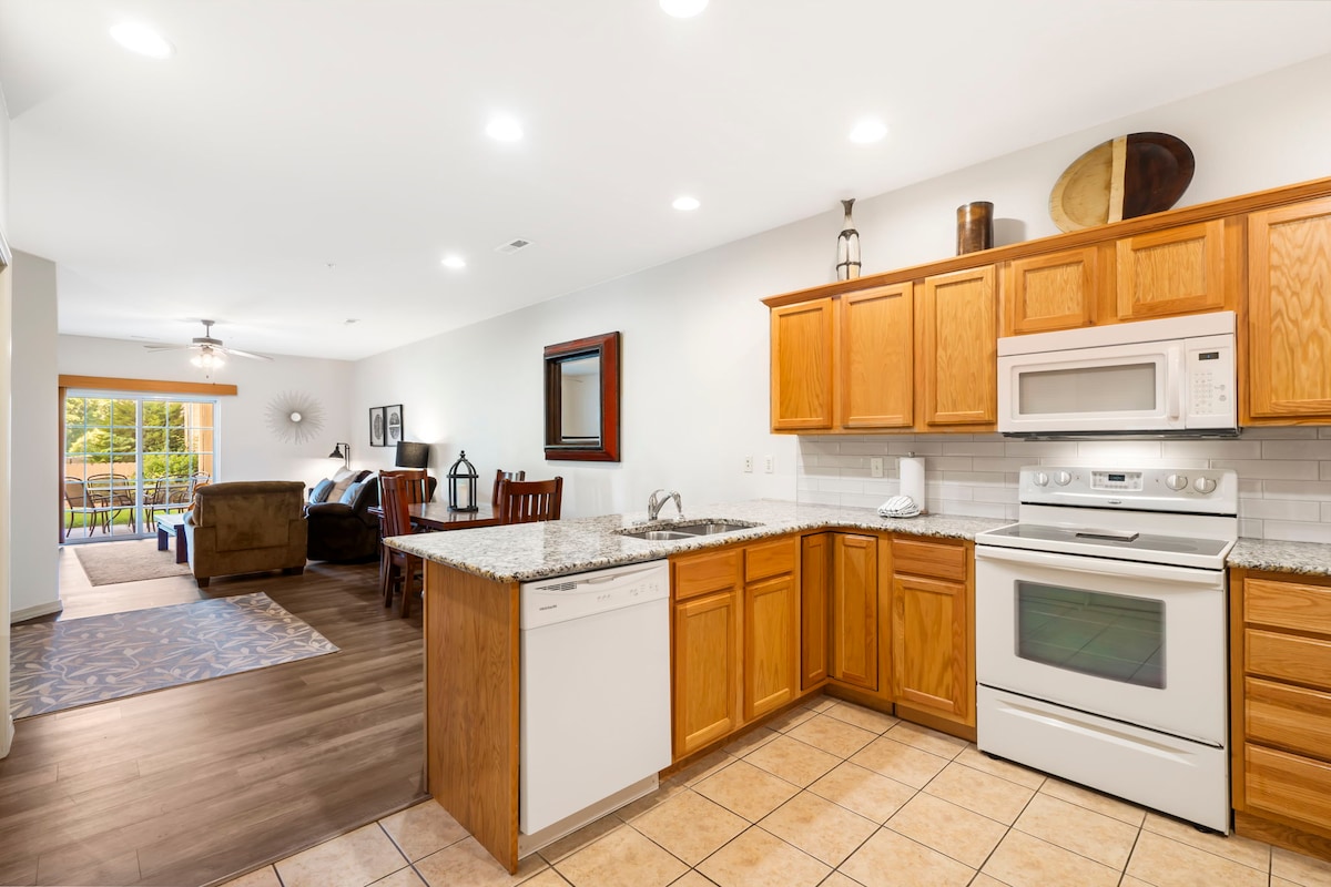 A well-equipped kitchen features light wood cabinetry and a granite countertop with a double bowl sink. Stainless steel appliances include a microwave and oven. An open layout leads to the dining area and living space, which is visible through a doorway, enhancing the connection between the two zones.