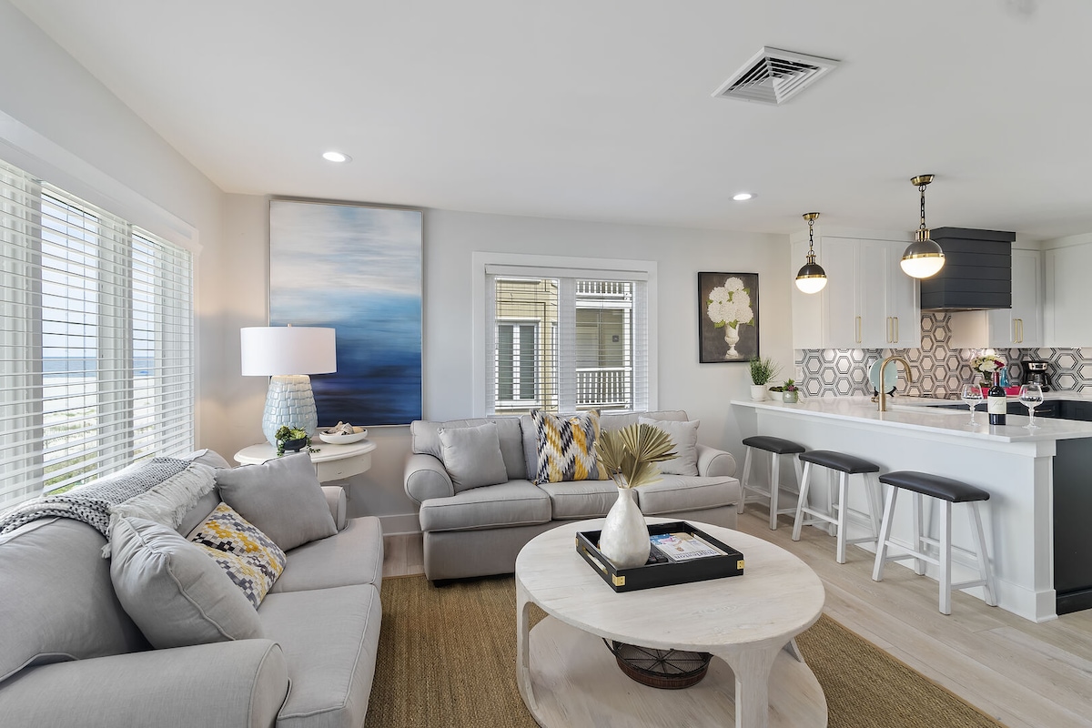 The living area features soft gray sofas and a round coffee table, complemented by natural light from multiple windows. A large piece of wall art with a blue theme enhances the decor, while sleek bar stools surround the modern kitchen's breakfast bar.