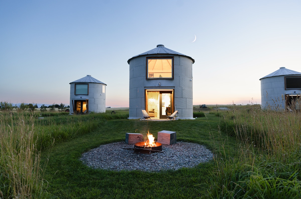 Two metal silos are positioned in a grassy area, each featuring large windows that reflect the dusk light. A circular fire pit with stone seating is centered in front, surrounded by lush grass, creating a welcoming outdoor space for relaxation.