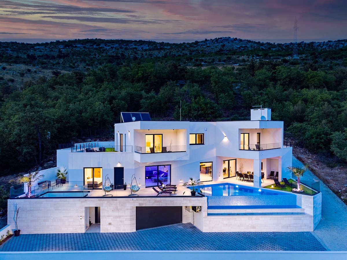 The villa is shown from an elevated perspective, highlighting its modern architecture and spacious layout. The infinity pool reflects the evening sky, while multiple glass doors provide access to indoor spaces. Lush greenery surrounds the property, enhancing the tranquil environment.