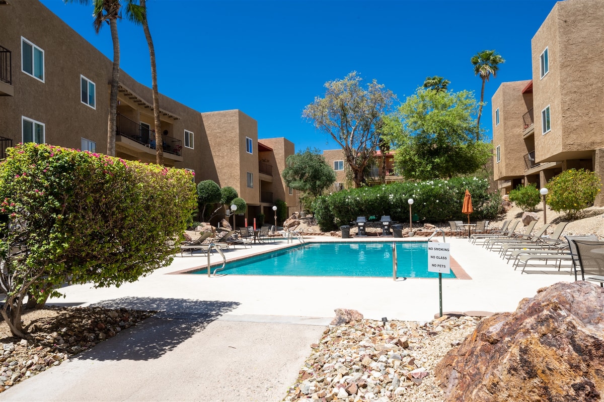 A community pool area is surrounded by landscaped greenery and patios. Lounge chairs are arranged along the poolside, and a few umbrellas provide shade. The clear blue sky complements the inviting setting, enhancing the space for relaxation and enjoyment.