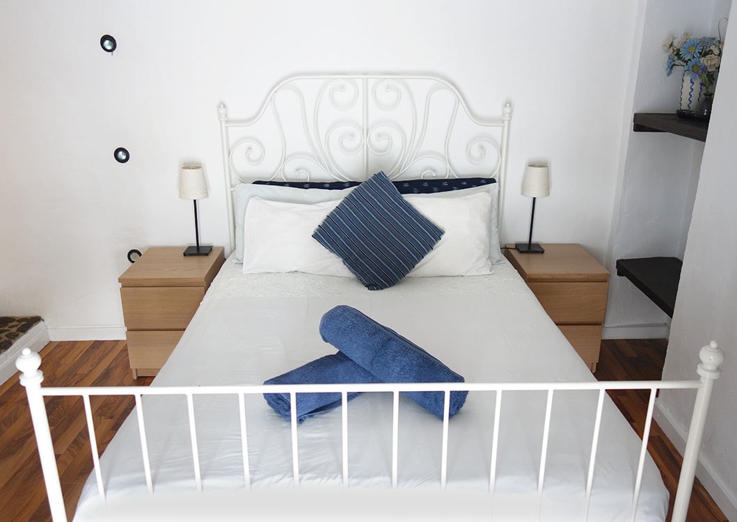 A private room features a double bed with a decorative headboard, complemented by two bedside tables equipped with lamps. Blue towels are neatly arranged on the bed, while a decorative pillow adds a touch of color. Wooden flooring enhances the room's design.