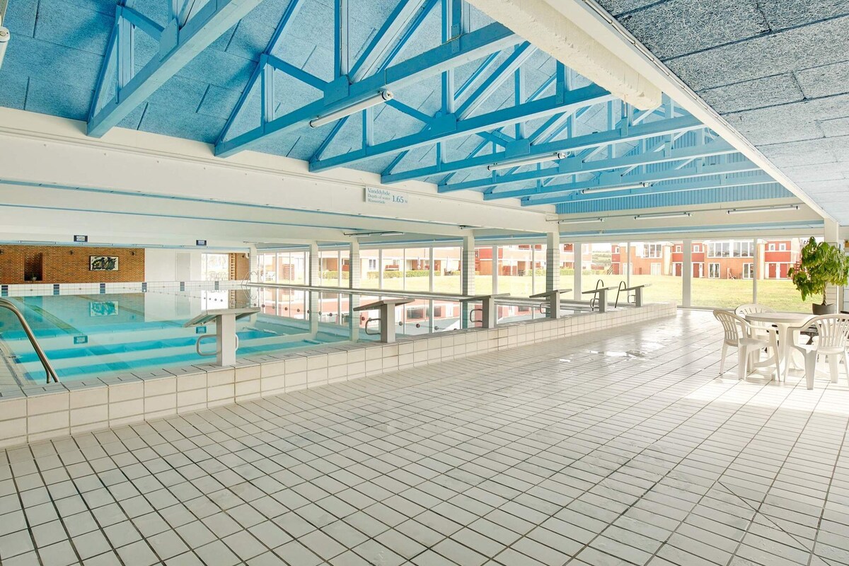 An indoor pool area is visible, featuring a large swimming pool with a shallow section. The ceiling is supported by blue beams, and the tiled floor contributes to a spacious environment. Nearby, a table and chairs are arranged for relaxation, with views toward the outdoor landscape.