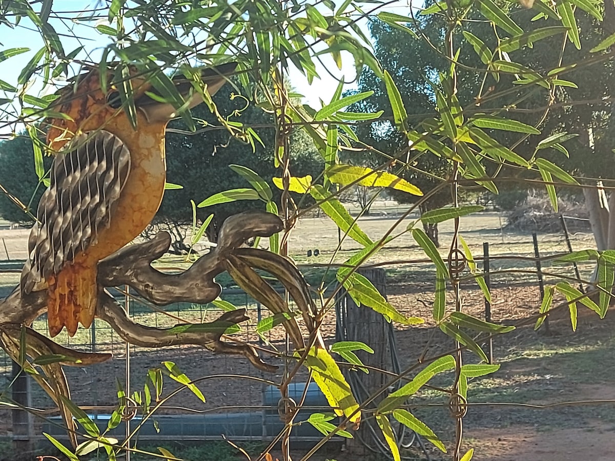 A decorative bird sculpture is perched amidst green foliage, providing a focal point against a natural background. Soft sunlight filters through the leaves, illuminating the scene, while a glimpse of the countryside is visible in the distance.