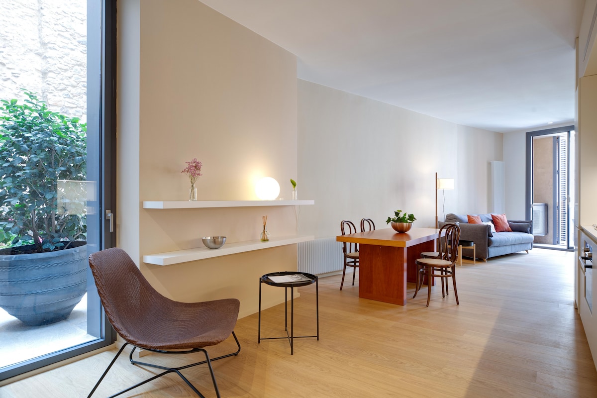 A modern living area features a cozy seating space with a sofa and a complementing coffee table. Natural light fills the room through large windows, highlighting the warm wooden flooring. Decorative plants and minimalist furnishings are present, creating a pleasant and serene environment.