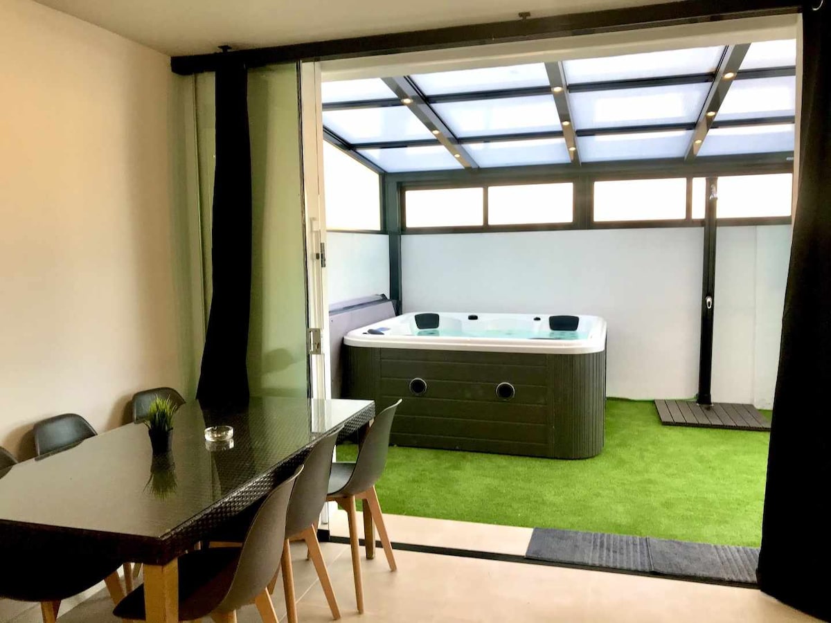 The image shows a modern indoor space overlooking a large jacuzzi, situated under a glass-roofed pergola. A dining table with black chairs is visible, complemented by artificial grass flooring, creating an inviting area for relaxation and socializing.