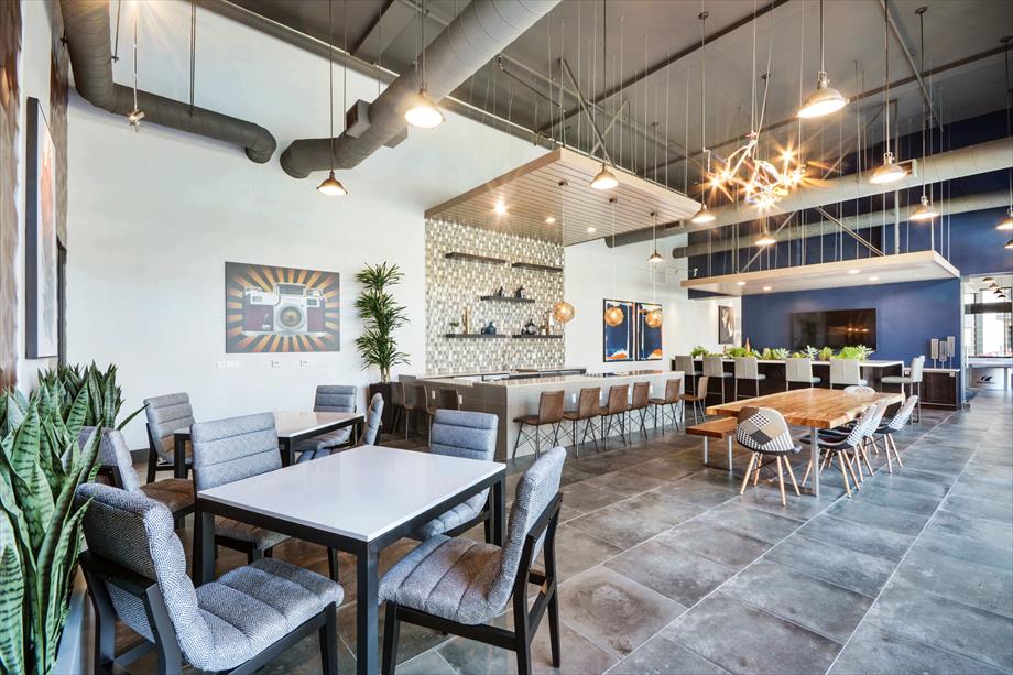 A modern communal lounge area is presented, featuring a mix of tables and seating arrangements. High ceilings are complemented by industrial-style light fixtures. A patterned feature wall adds visual interest, while greenery is incorporated into the space, creating an inviting environment for relaxation and socializing.