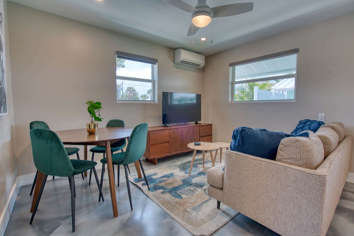 A stylish living area is furnished with a comfortable sofa and a circular coffee table. A dining table with four green chairs complements the space. Natural light is provided by two windows, and a flat-screen television is positioned on a wooden media console across the room.