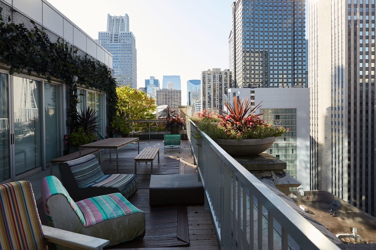 An outdoor rooftop terrace features a variety of seating, including a striped lounge chair and wooden tables. Lush greenery surrounds the area, providing a tranquil space. The skyline of downtown Chicago, characterized by tall buildings and vibrant architecture, is visible in the background.