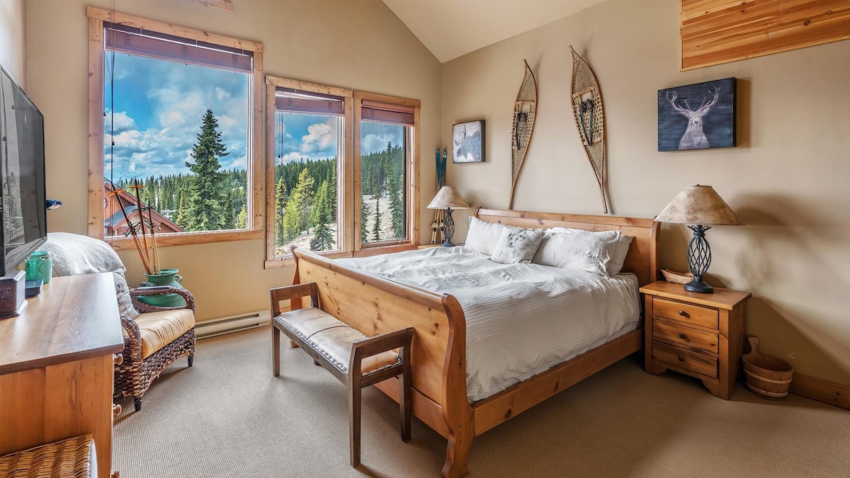 A serene bedroom presents natural wood furnishings, including a spacious bed with white linens. Large windows offer expansive views of the surrounding trees and mountains. Accents include decorative wall art and a cozy seating area with a woven chair.