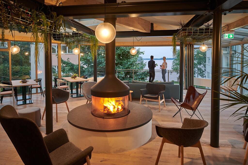 A modern lounge area is centered around a circular fireplace, surrounded by comfortable seating. Large windows offer views of the outdoors, where two individuals are seen enjoying the scenery. Pendant lighting creates a warm ambiance, complemented by indoor plants adorning the space.