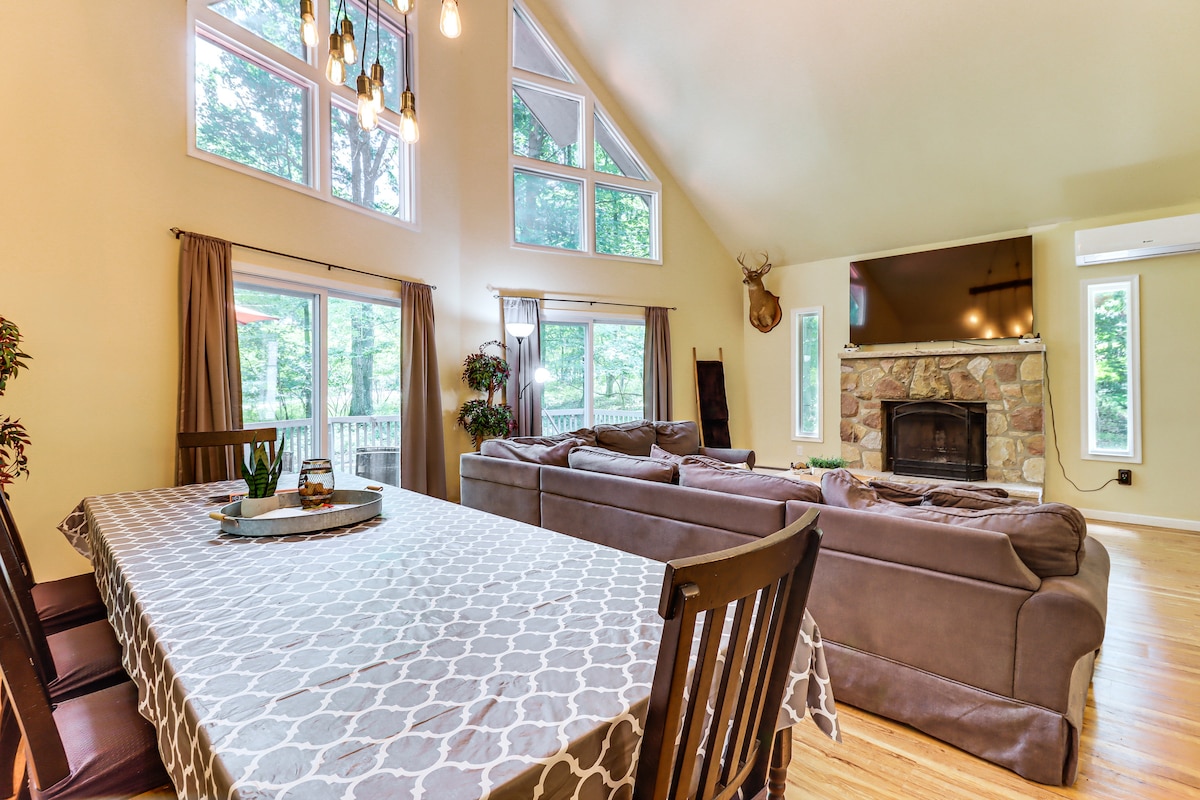 A bright and airy living space features a high ceiling with large windows allowing natural light to fill the area. A seating arrangement faces a stone fireplace, while a dining table is set with a decorative centerpiece, inviting shared meals and gatherings.
