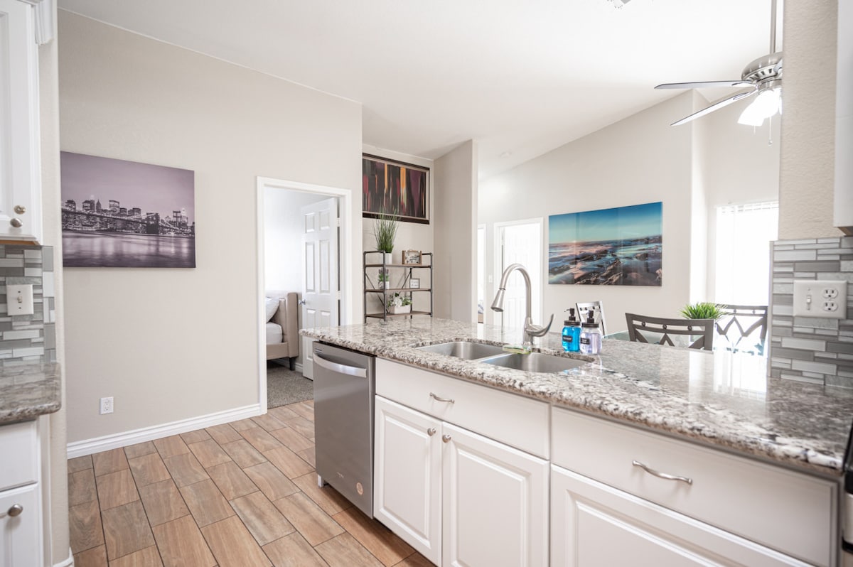 A modern kitchen displays sleek granite countertops and white cabinets. Stainless steel appliances, including a dishwasher, contribute to the functional design. Natural light enters from nearby windows, illuminating the space, while decorative art pieces provide a touch of color to the neutral walls.