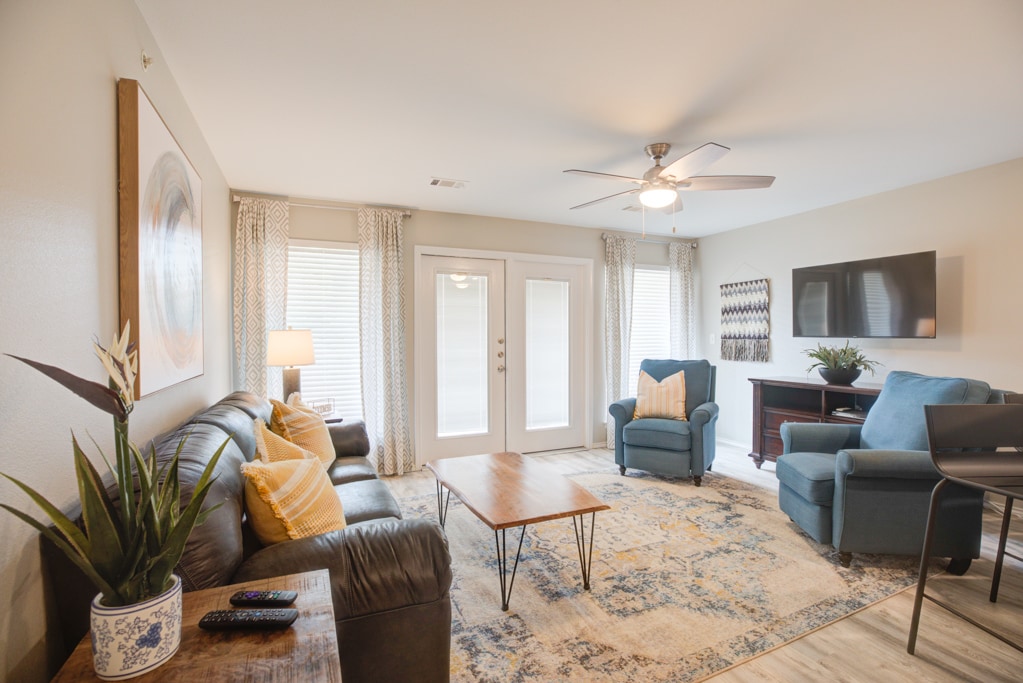 A comfortable living area is featured, highlighted by a leather sofa and blue armchairs. A wooden coffee table sits on a patterned area rug. Natural light filters through the windows, and a television is mounted on the wall opposite, creating a relaxed atmosphere.