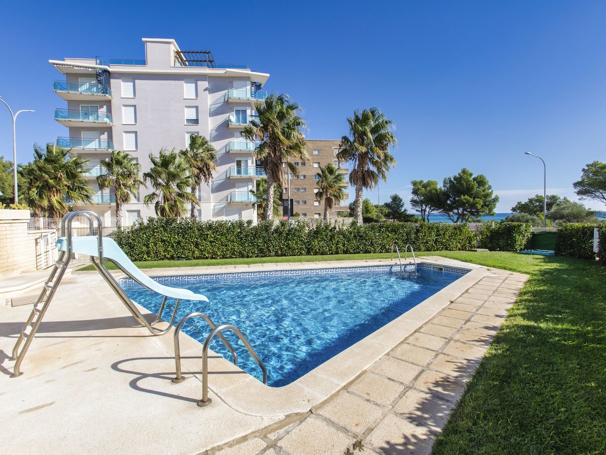 A well-maintained swimming pool features a gentle slope and slides into the clear water. Lush greenery surrounds the area, with palm trees providing shade. The distant view reveals a glimpse of the sea, complemented by a clear blue sky.