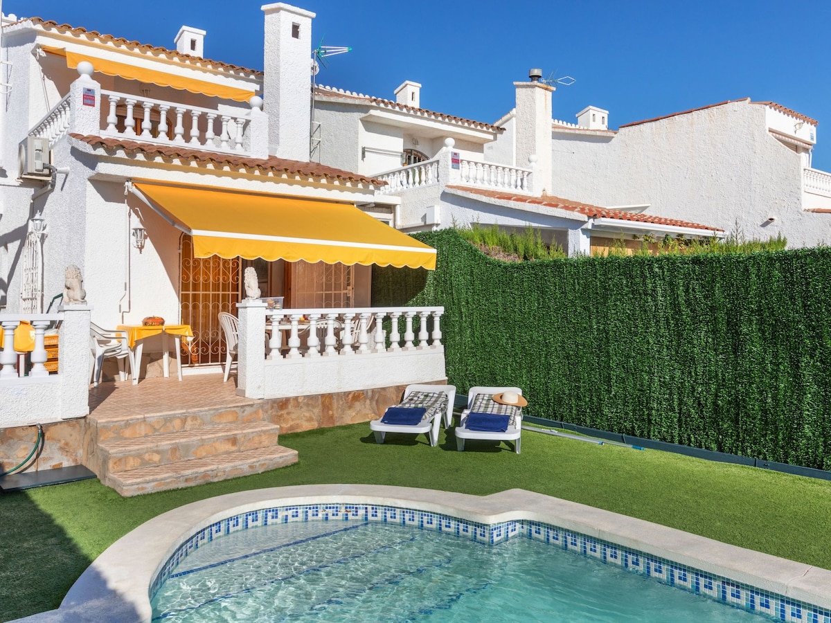 A sunny terrace features a yellow awning over a dining area, adjacent to a private garden and a swimming pool. Sun loungers are positioned beside the pool, surrounded by manicured greenery. The white façade of the two-story house reflects a welcoming atmosphere.