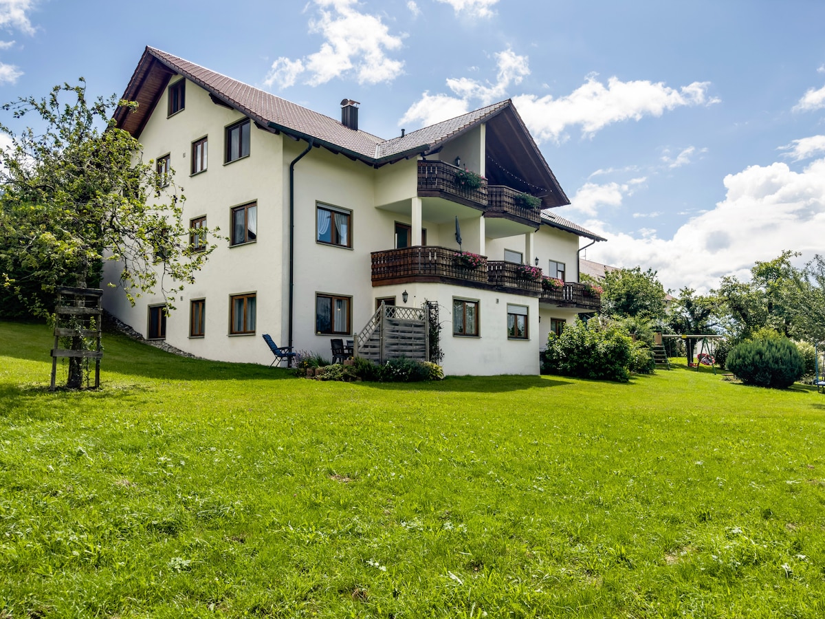 The exterior of a three-story farmhouse is displayed, featuring a sloped roof and multiple balconies. A spacious lawn surrounds the building, with trees and greenery enhancing the landscape. Sunlight brightens the scene, and the sky is partly cloudy, contributing to a serene atmosphere.