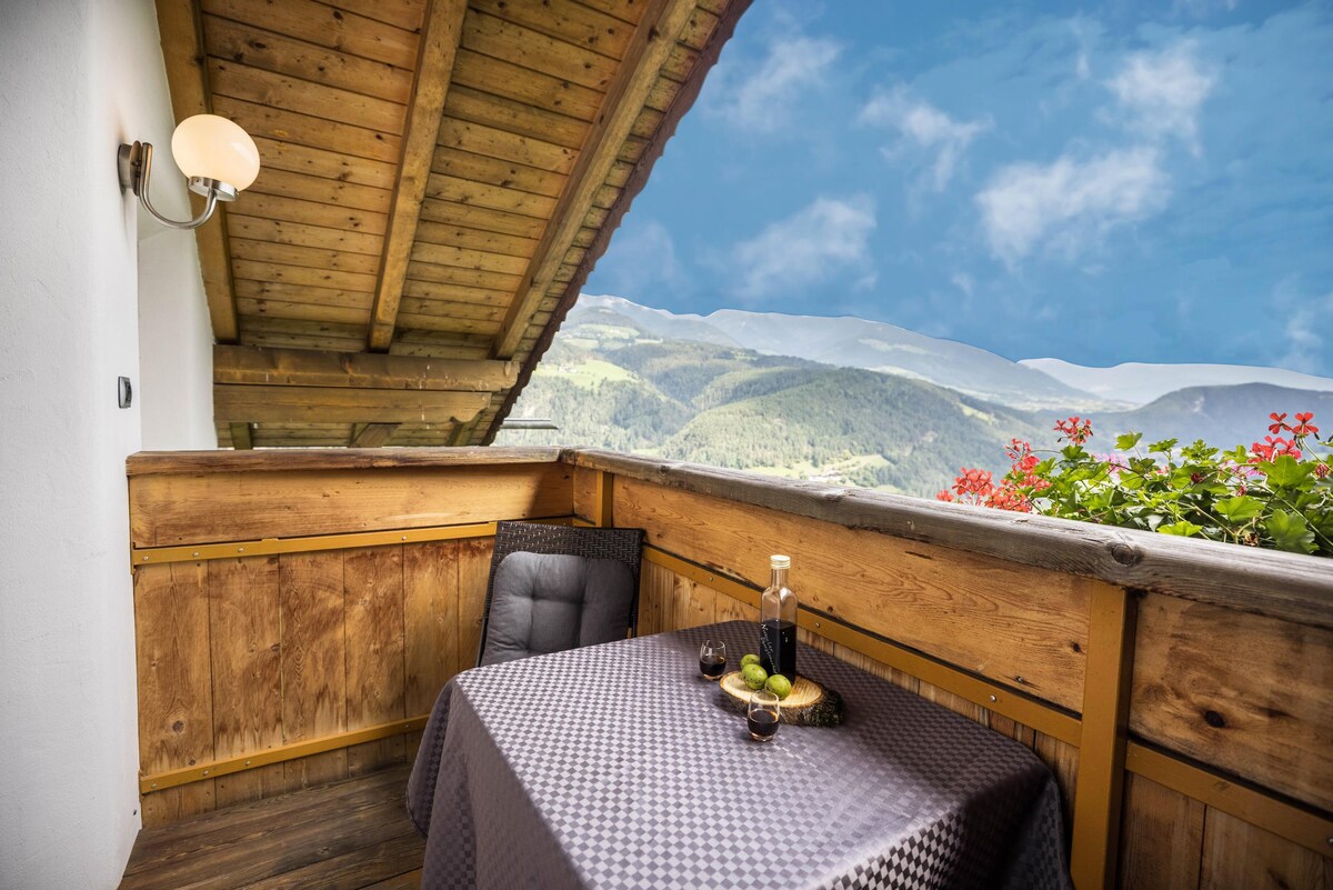 A cozy balcony is presented with wooden railings and a table covered with a patterned cloth. A bottle and glasses are placed on the table, surrounded by lush greenery and vibrant flowers. Scenic mountain views stretch into the distance under a clear blue sky.