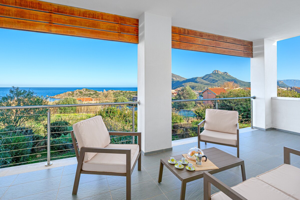 A covered terrace offers scenic views of the ocean and surrounding hills. Two light-colored armchairs and a small table are positioned on the tile floor, inviting relaxation. Natural light brightens the space through large windows that provide an open, airy feel.