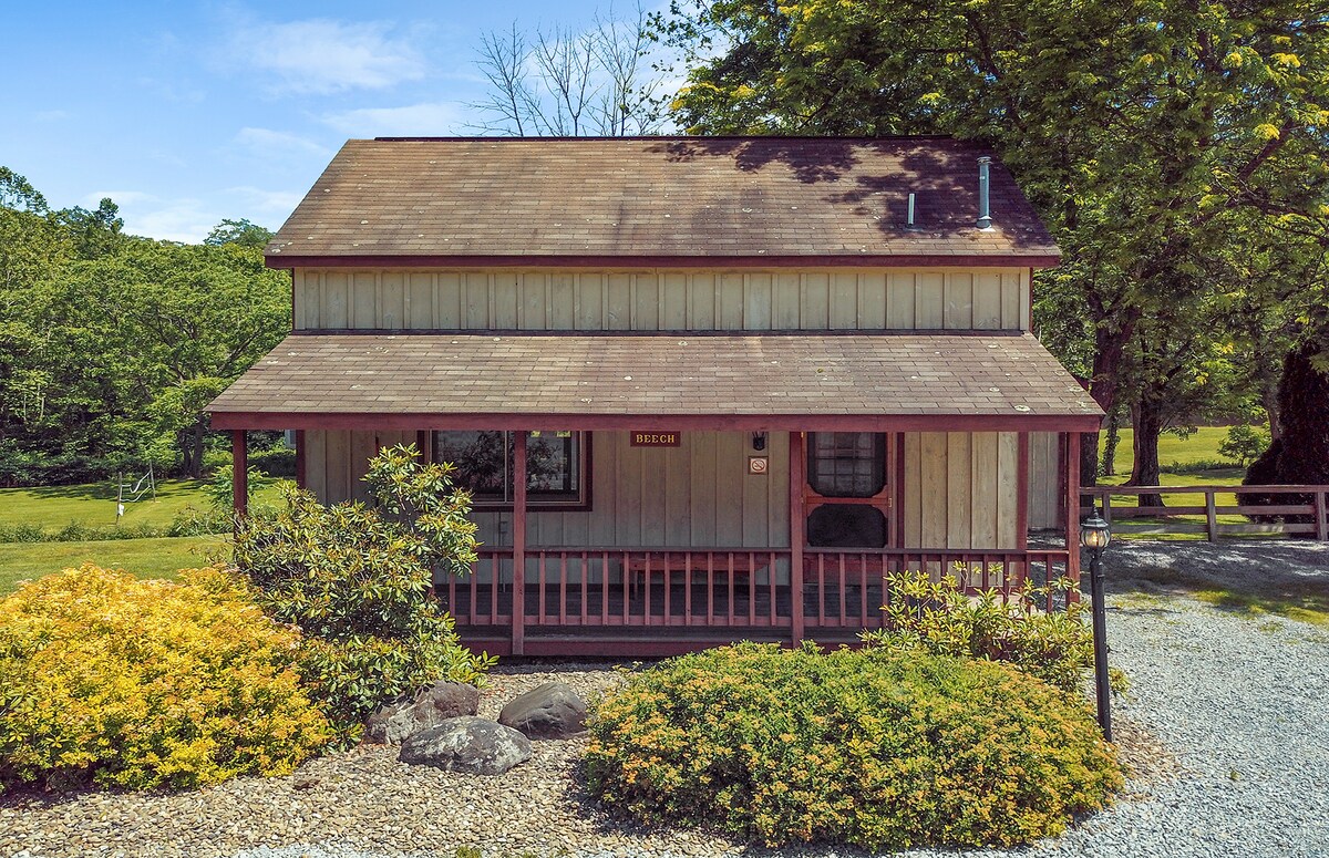 The two-story cabin is framed by a porch featuring a railing and a mix of greenery and landscaping. The exterior showcases wooden siding and a gabled roof, with large windows allowing natural light to enter. Surrounding nature adds to the serene environment.
