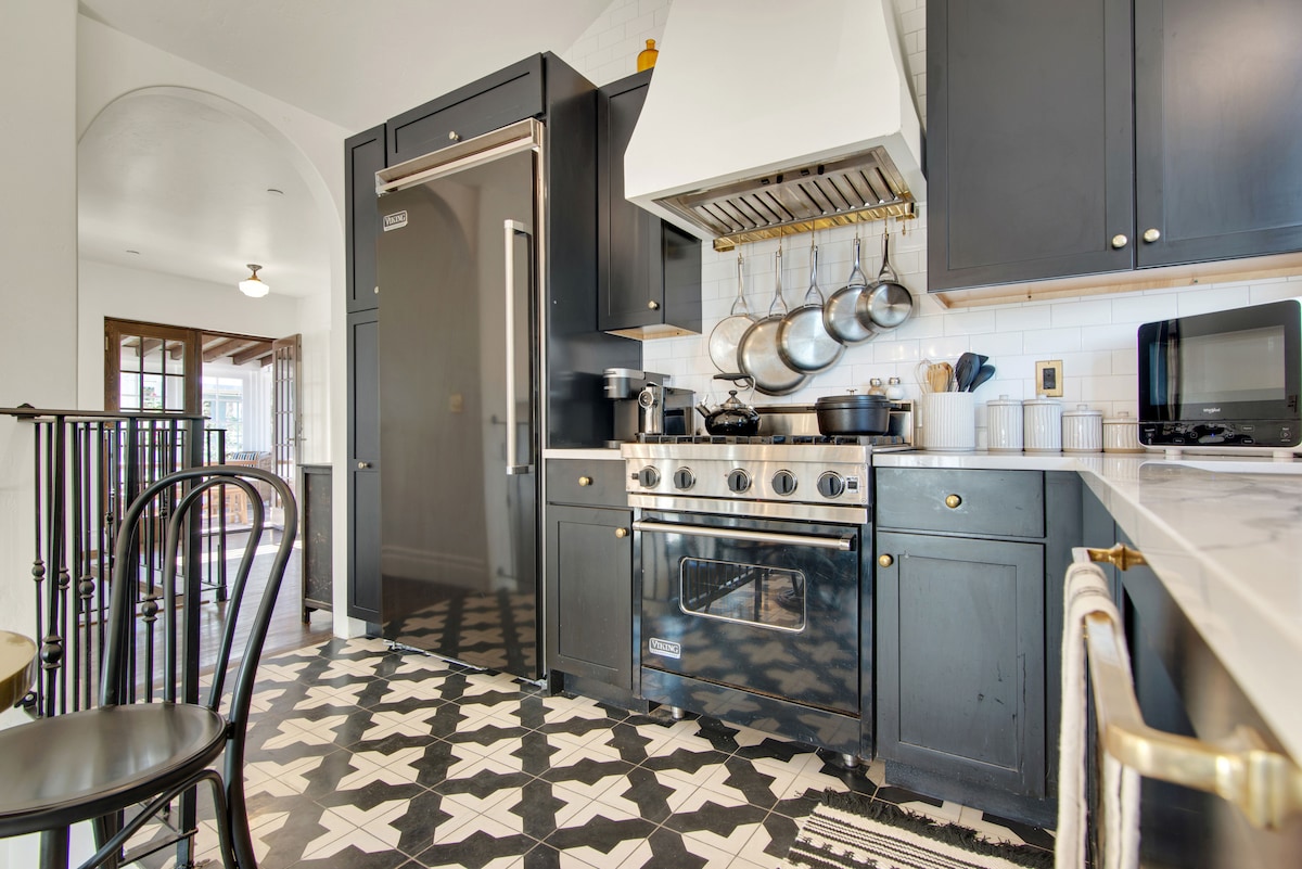 The kitchen features black cabinetry and a stainless steel stove with a vent hood above. Hanging cookware is present, and a sleek refrigerator is nearby. A tiled floor with a geometric pattern enhances the modern aesthetic, while ample natural light fills the space.