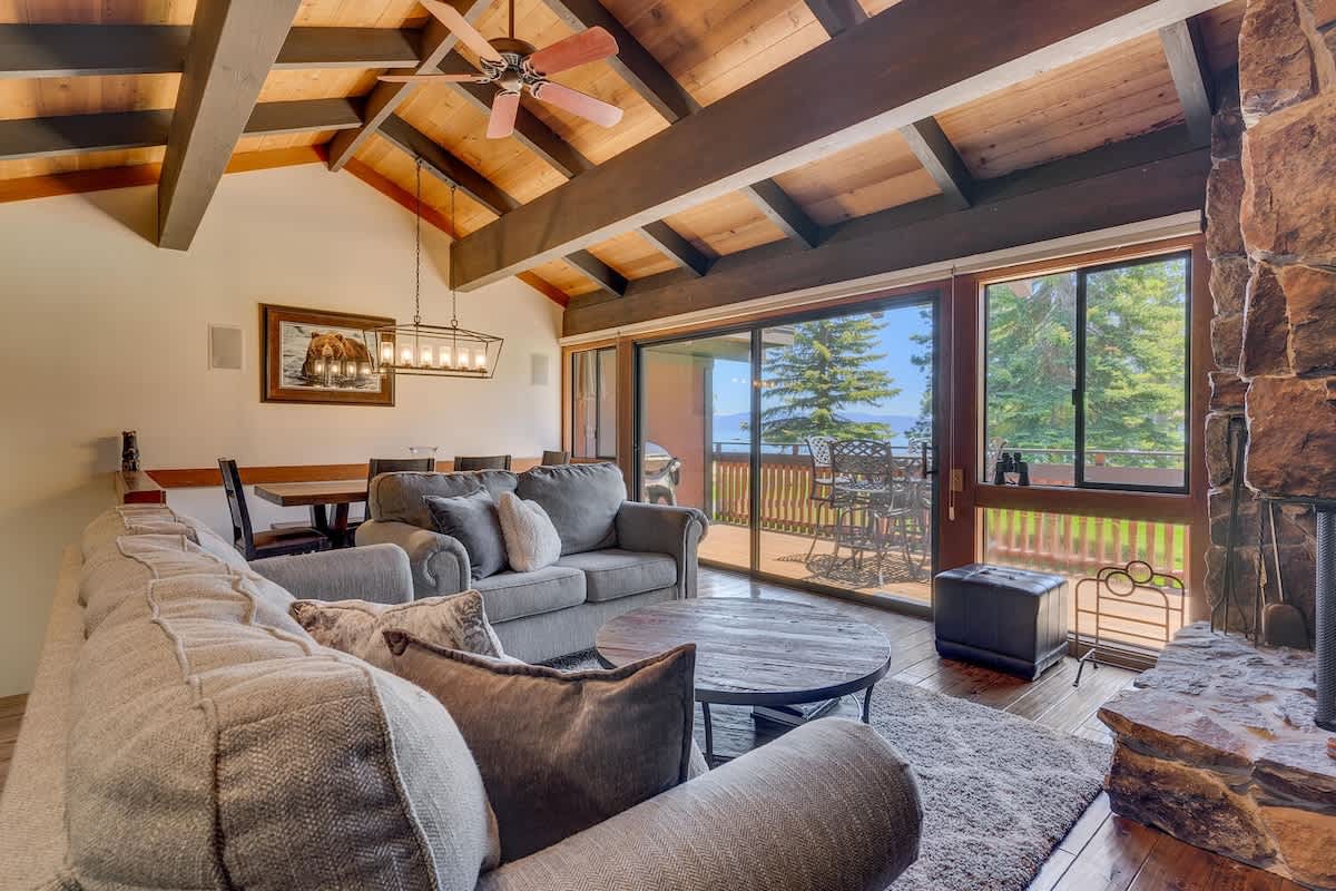 An inviting living room features a spacious sectional sofa arranged around a circular coffee table. Large glass doors provide views of the outdoor deck, and wooden beams accentuate the high ceiling. A dining area with a chandelier is visible in the background.