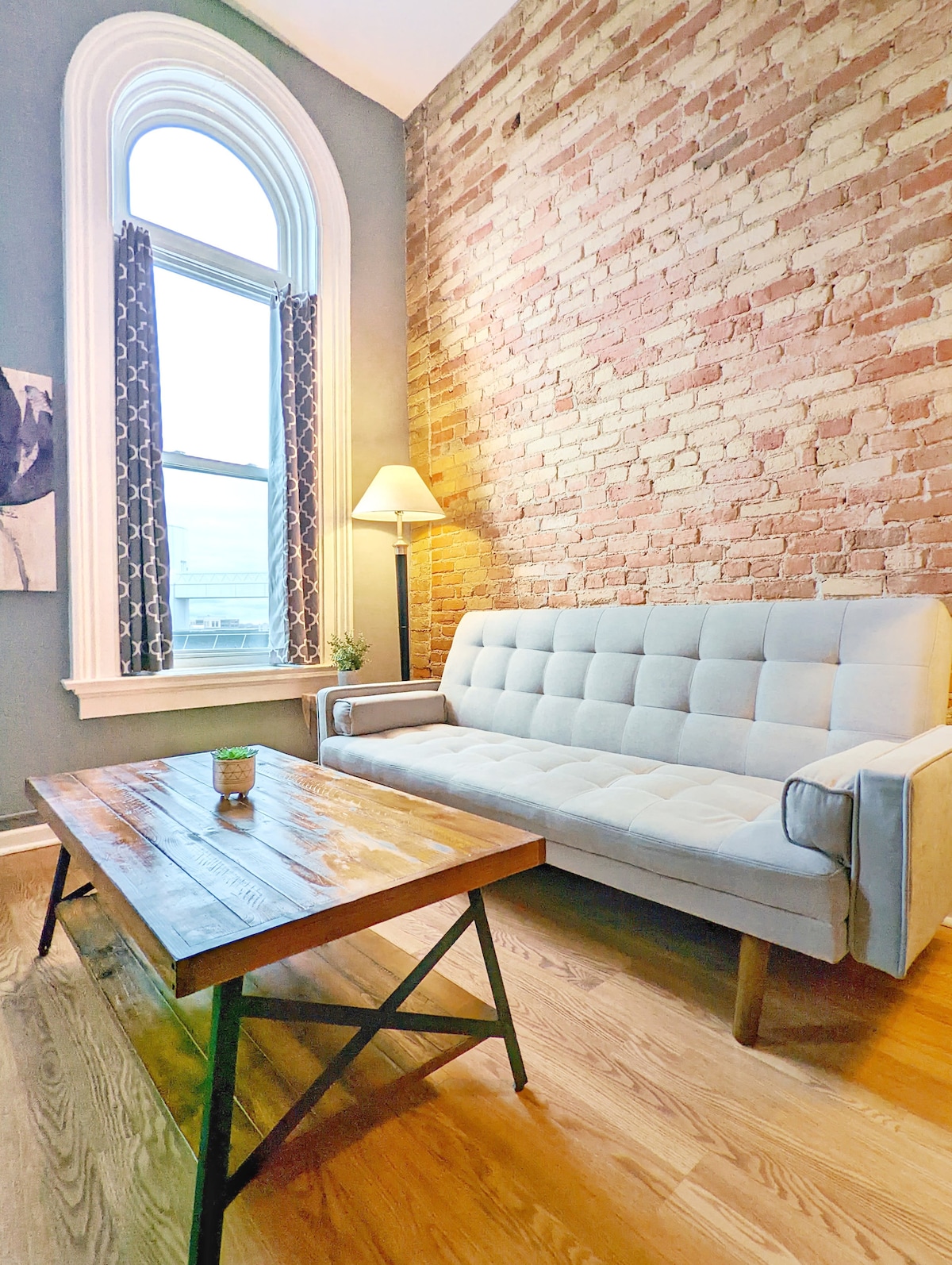 A light-filled living area features a comfortable sofa against a brick wall, complemented by a wooden coffee table. Large arched windows allow natural light to illuminate the space, while patterned curtains frame the view.