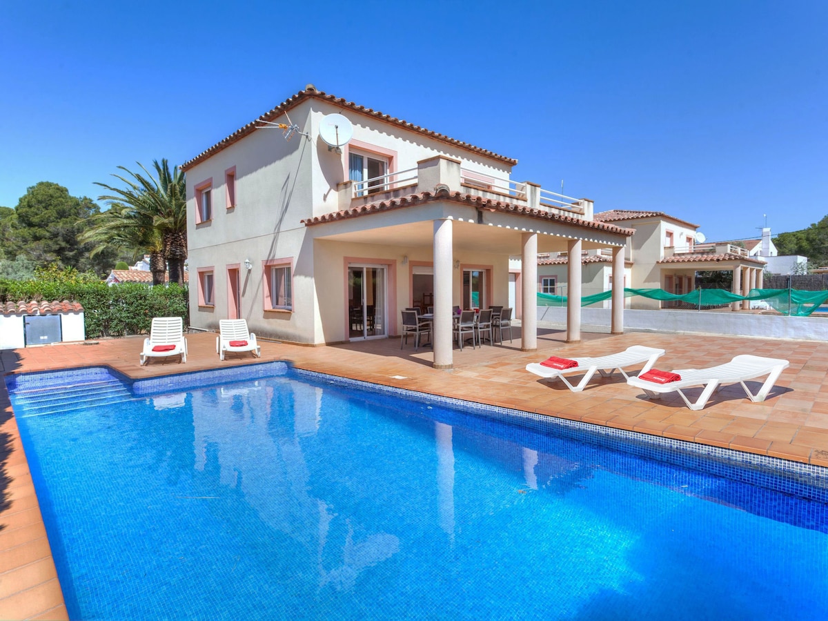 The two-story villa is shown with a spacious terrace and private swimming pool. Sun loungers are arranged around the pool, with a dining table visible on the terrace. Lush greenery surrounds the property, providing a serene atmosphere.
