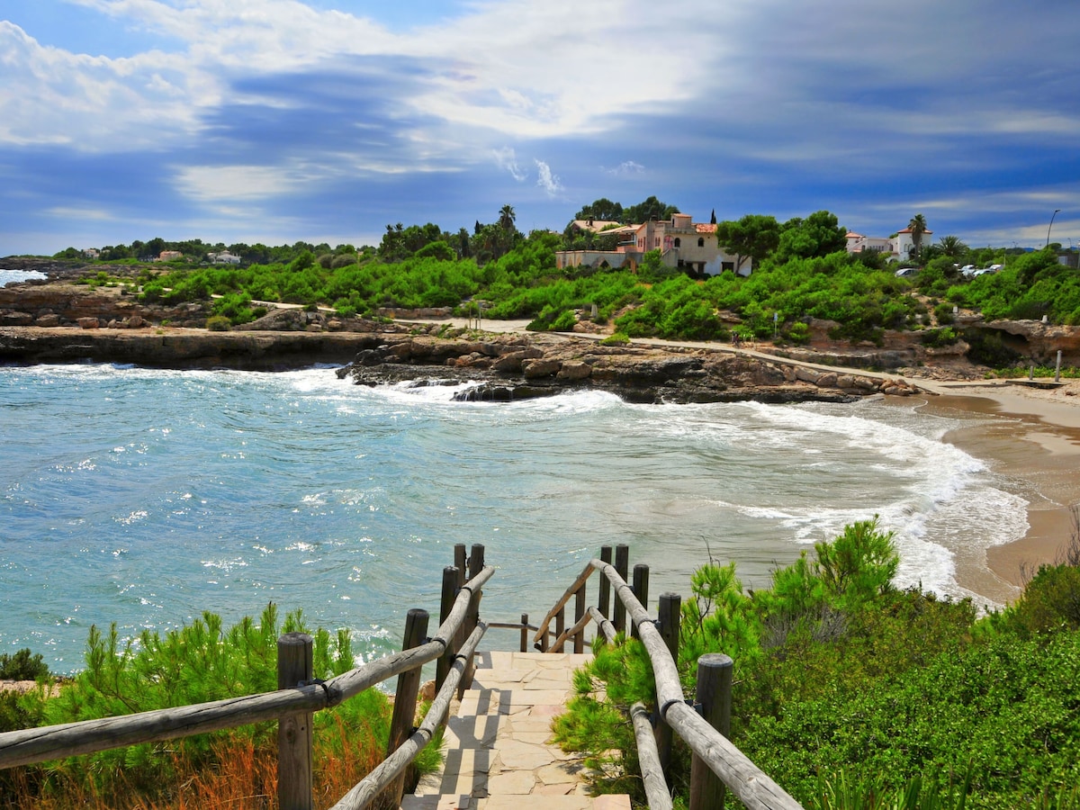 A scenic pathway leads down to a tranquil beach, flanked by lush greenery and rocky formations. The gentle waves lap at the shoreline under a cloudy sky, with a coastal villa visible in the background amidst the vegetation.