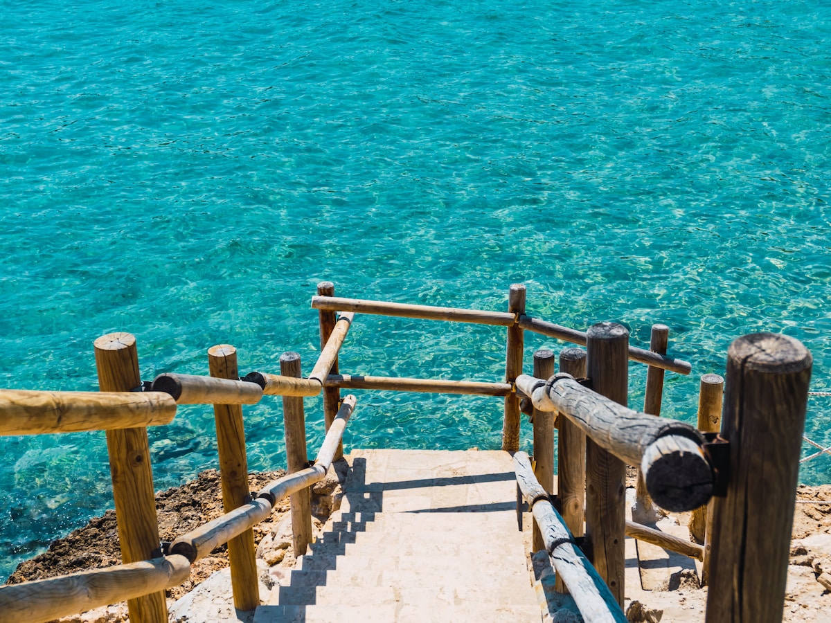 A wooden staircase descends towards clear blue waters, surrounded by a rustic railing. The vibrant turquoise sea reflects sunlight, creating a serene and inviting atmosphere. The rocks lining the shore are visible, blending naturally with the coastal landscape.