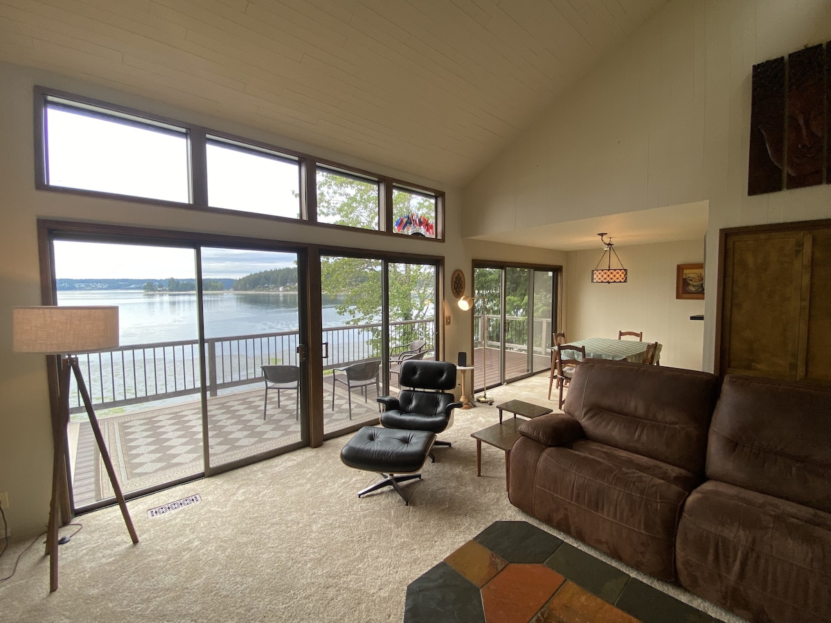 A spacious living area features large glass doors that open to panoramic water views. A dark recliner and a plush couch are positioned for comfort, while a dining space with a table and chairs is visible nearby. Natural light floods the space through the windows.