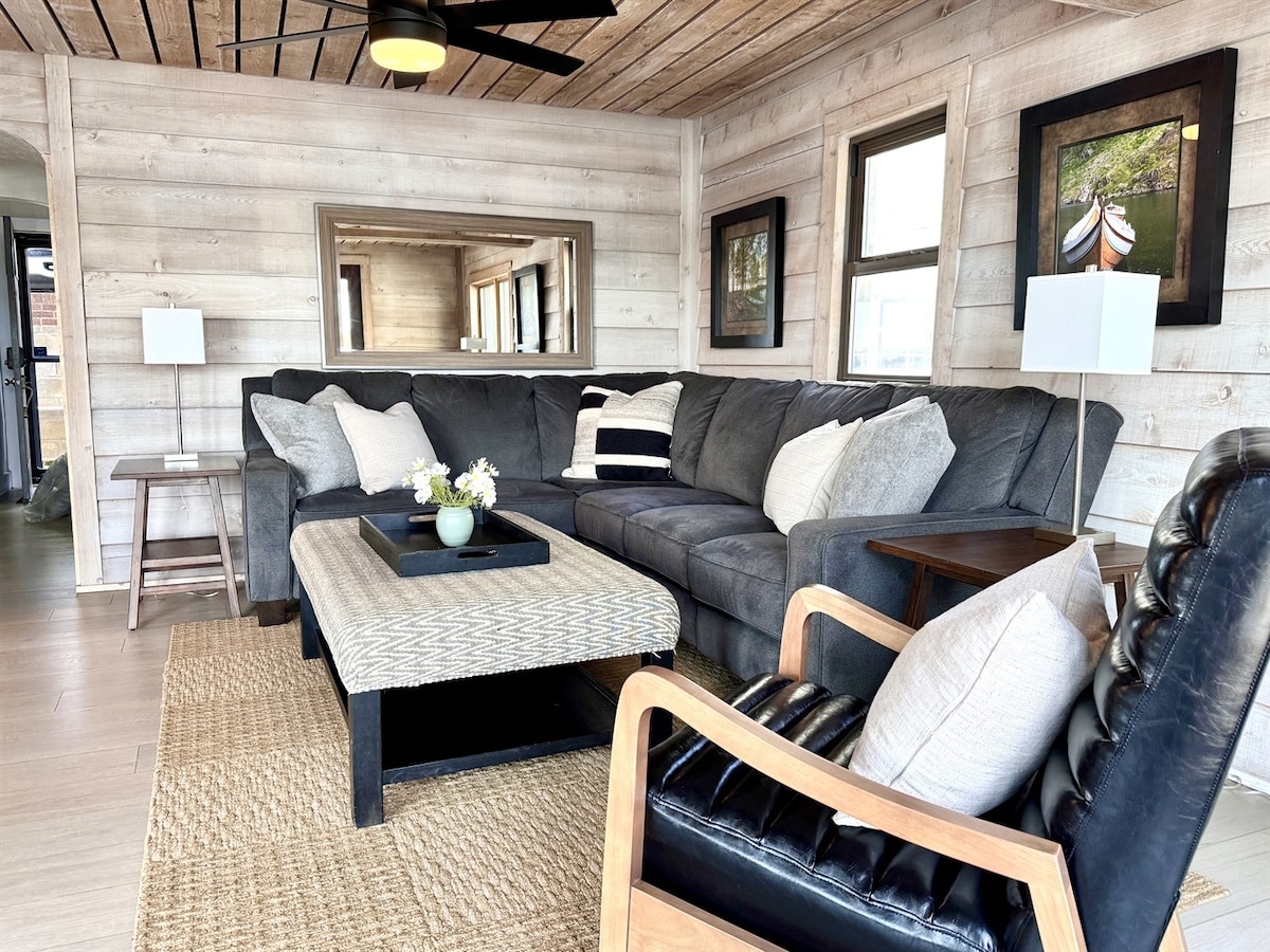 A spacious living area features comfortable seating with a large sectional sofa, complemented by a textured area rug. Soft lighting enhances the inviting atmosphere, while large windows provide natural light and views of the outdoors. A coffee table and side chairs offer additional seating options.