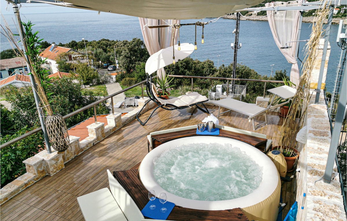 An outdoor terrace features a round jacuzzi surrounded by wooden decking, with lounge chairs positioned for sea views. The space is adorned with draped fabric overhead and offers a sense of openness, inviting relaxation while taking in the scenic coastal landscape.